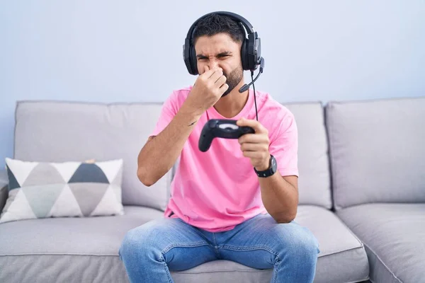 西班牙裔年轻人坐在沙发上玩电子游戏 手里拿着控制器 闻到一股难闻难闻的气味 用手指捂住鼻子屏住呼吸 — 图库照片