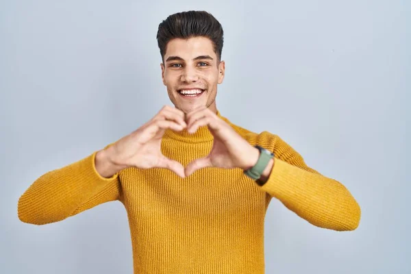青い背景の上に立つ若いヒスパニック系の男が手でハートのシンボル形状を行う愛に笑みを浮かべて ロマンチックなコンセプト — ストック写真
