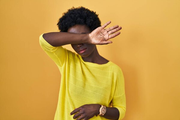 African young woman standing over yellow studio covering eyes with arm, looking serious and sad. sightless, hiding and rejection concept