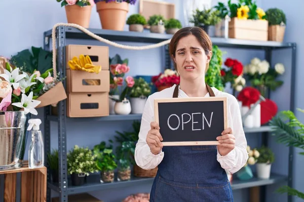 オープンサインを保持する花屋で働くブルネットの女性は混乱した表情 疑わしい概念 — ストック写真
