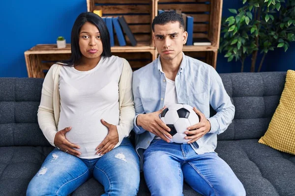 若いヒスパニック系のカップルは 赤ちゃんがソファにボールを保持し おなかが落ち込んで座って期待し 苦痛のために心配し 怒っていると恐れて泣いています 悲しい表情 — ストック写真