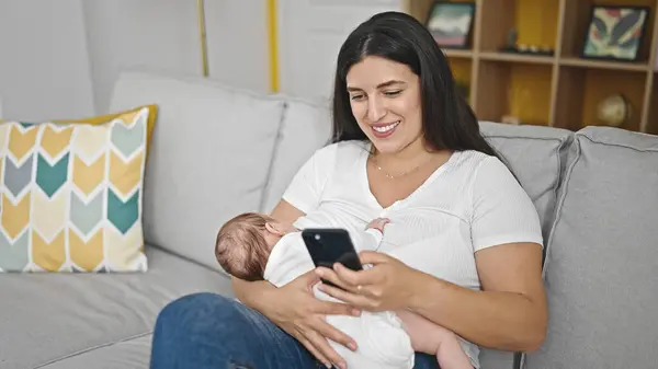 自宅でスマートフォンを使用してソファーの授乳中の赤ちゃんに座っている母と赤ちゃんの娘 — ストック写真