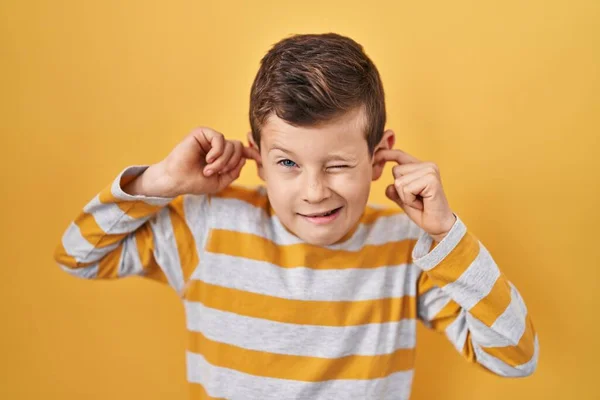 若い白人の子供は 大きな音楽のノイズのためにいらいらする表現で指で耳を覆う黄色の背景に立っています 聴覚障害の概念 — ストック写真