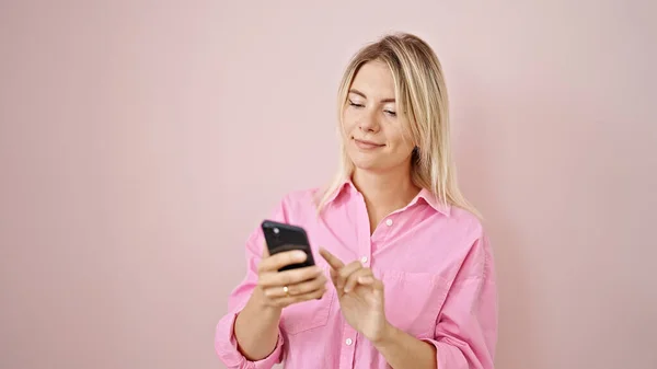 年轻的金发女人用智能手机对着孤立的粉色背景微笑 — 图库照片
