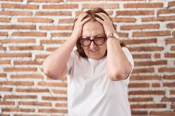 老年妇女戴着眼镜站在砖墙上 由于疼痛和偏头痛 绝望和压力重重 手放在头上 — 图库照片