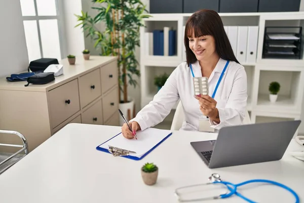 stock image Young beautiful hispanic woman doctor holding pills bottle writing on document at clinic