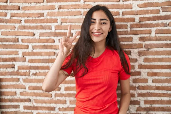年轻的少女站在砖墙上 微笑着看着摄像机 手指头在做胜利的标志 第二点 — 图库照片
