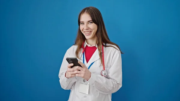 年轻的惊慌失措的女医生用智能手机对着孤立的蓝色背景微笑 — 图库照片