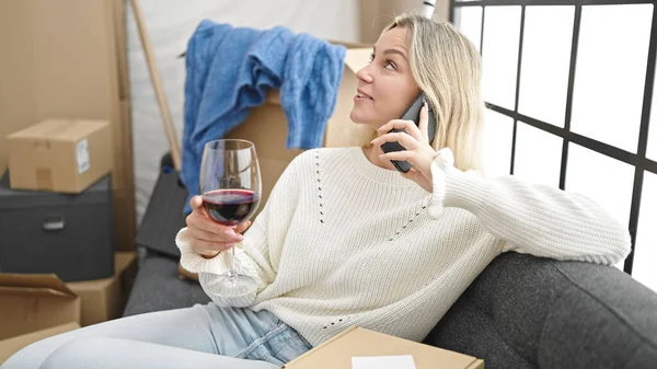 Young blonde woman talking on the smartphone and drinking wine at new home