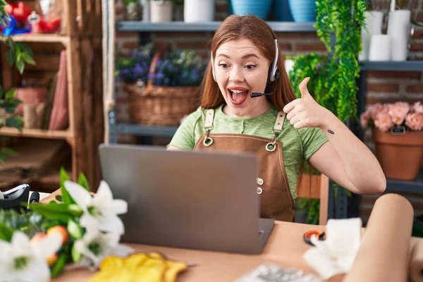 Redhead woman working at florist shop doing video call smiling happy and positive, thumb up doing excellent and approval sign