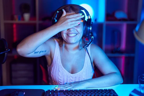 若いブロンドの女性は ヘッドフォンの笑顔を着て 驚きのために目を覆う顔に手で笑ってビデオゲームをしています 視覚障害の概念 — ストック写真
