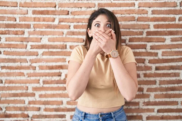レンガの壁の上に立って若いブルネットの女性は間違いのために手で口をカバーショックを受けた 秘密の概念 — ストック写真