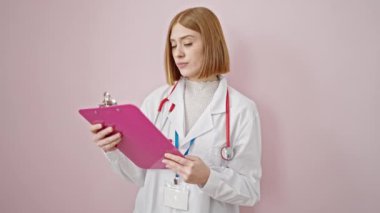 Genç sarışın kadın doktor, pembe arka planda not panosundan belge okuyor.