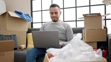 İspanyol adam yeni evinde dizüstü bilgisayar kullanarak gülümsüyor.
