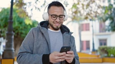 İspanyol adam, parkta akıllı telefon kullanarak müzik dinlerken kendinden emin gülümsüyor.