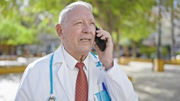 一位头发灰白的高级男医生站在公园里 严肃地在智能手机上讲话 — 图库视频影像