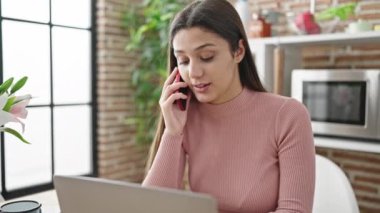 Genç ve güzel İspanyol kadın yemek odasında dizüstü bilgisayar kullanıyor ve akıllı telefondan konuşuyor.