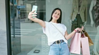Genç ve güzel İspanyol bir kadın elbise mağazasında elinde çantalarla akıllı telefonuyla selfie çekiyor.