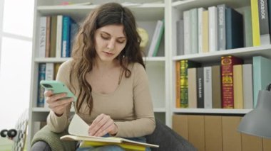 Genç ve güzel İspanyol kız öğrenci kütüphanede akıllı telefon okuma kitabı kullanıyor.