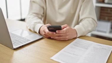 Genç beyaz adam yemek odasında dizüstü bilgisayar ve akıllı telefon kullanıyor.