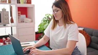 Laptop kullanan genç sarışın kadın yemek odasında gülümseyen gözlükleri çıkarıyor.