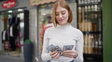Young blonde woman smiling confident counting dollars at street