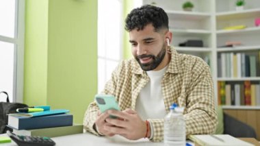 Genç İspanyol öğrenci kütüphanede akıllı telefon ve kulaklık kullanıyor.