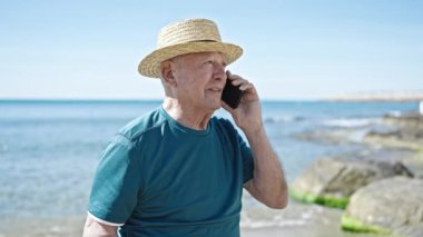 Kıdemli kır saçlı turist. Yazlık şapka takıyor. Deniz kenarında akıllı telefondan konuşuyor.