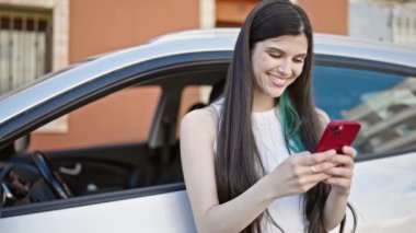 Young beautiful hispanic woman using smartphone leaning on car at street