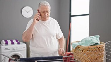 Orta yaşlı, kır saçlı, akıllı telefondan konuşan çamaşır odasında çamaşır ipine kıyafet asan bir adam.