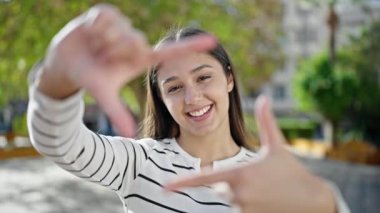Genç, güzel, İspanyol bir kadın gülümsüyor. Parkta elleri ile çerçeve hareketi yapıyor.