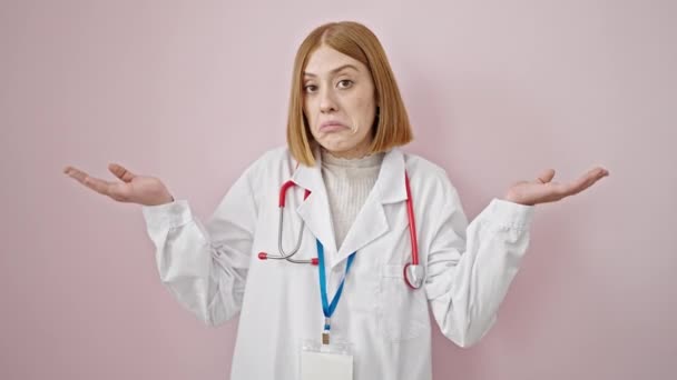 若いですブロンド女性医師立っていますClueless上の隔離されたピンクの背景 — ストック動画
