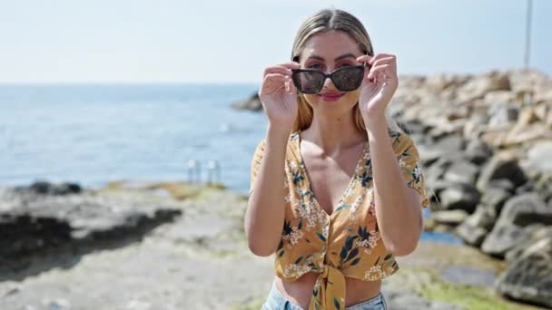 Młoda Blondynka Uśmiecha Się Pewnie Nosząc Okulary Przeciwsłoneczne Nad Morzem — Wideo stockowe