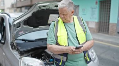 Orta yaşlı, kır saçlı bir adam araba arızası için akıllı telefondan sigortaya mesaj atıyor.