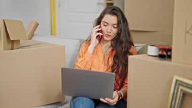 Genç, güzel, İspanyol bir kadın yeni evinde bilgisayarla akıllı telefondan konuşuyor.