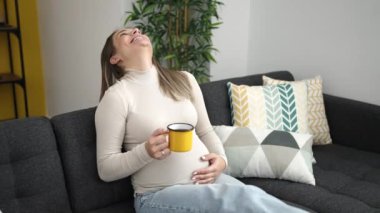 Genç hamile kadın kahvesini yudumlarken karnına dokunuyor.