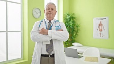 Orta yaşlı, gri saçlı, gülümseyen bir doktor klinikte kollarını kavuşturup el kol hareketi yapıyor.