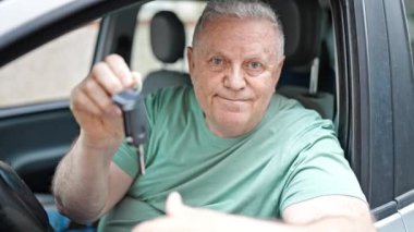 Orta yaşlı, kır saçlı bir adam yeni arabanın anahtarını tutuyor. Sokakta kazanan yüz ifadesi var.
