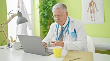 Orta yaşlı, gri saçlı bir adam. Laptop kullanıyor. Klinikte kahve içiyor.
