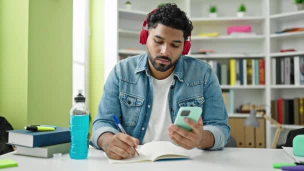 アフリカ系アメリカ人の学生が図書館大学でスマートフォンやヘッドフォンを使ってノートを書く — ストック動画