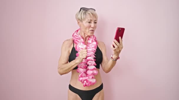 中年金发女人穿着比基尼和夏威夷莱雅 带着独立的粉色背景的视频通话 — 图库视频影像