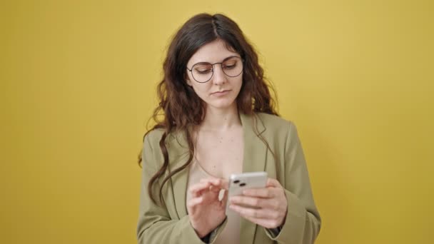 年轻美丽的惊慌失措的女人用智能手机对着孤立的黄色背景微笑 — 图库视频影像