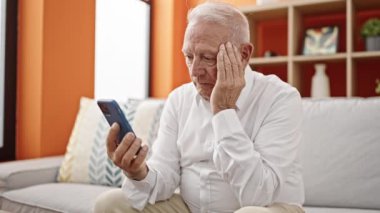 Yaşlı gri saçlı adam evde akıllı telefon kullanmaktan endişeleniyor.