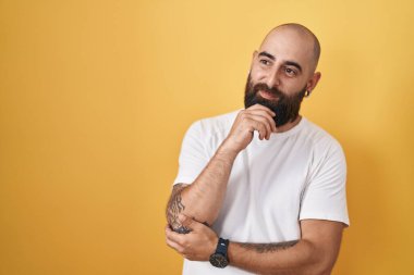 Sakallı ve dövmeli genç İspanyol bir adam. Sarı arka planda duruyor. Elleri çenesinde soru sormayı düşünüyor. Düşünceli bir ifade. Gülümseyen ve düşünceli bir yüz. şüphe kavramı. 