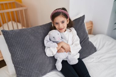 Sevimli İspanyol kız yatakta oturmuş oyuncak bebeğe sarılıyor.