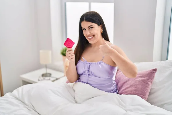 stock image Young brunette woman wearing lingerie and holding condom on the bed smiling happy and positive, thumb up doing excellent and approval sign 