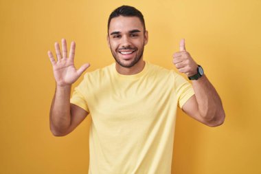 Sarı arka planda dikilen İspanyol genç adam altı numaralı parmaklarıyla gülümseyerek kendinden emin ve mutlu bir şekilde işaret ediyor.. 