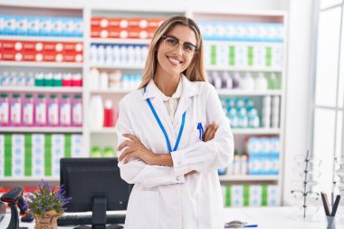 Young blonde woman pharmacist smiling confident standing with arms crossed gesture at pharmacy