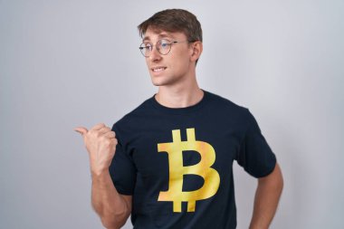 Beyaz, sarışın, bitcoin tişörtlü, gülen yüzlü bir adam başparmağıyla yan tarafa bakıyor ve gülümsüyor.. 