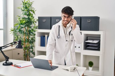 Doktor üniforması giyen İspanyol genç adam klinikte dizüstü bilgisayarla telefonla konuşuyor.